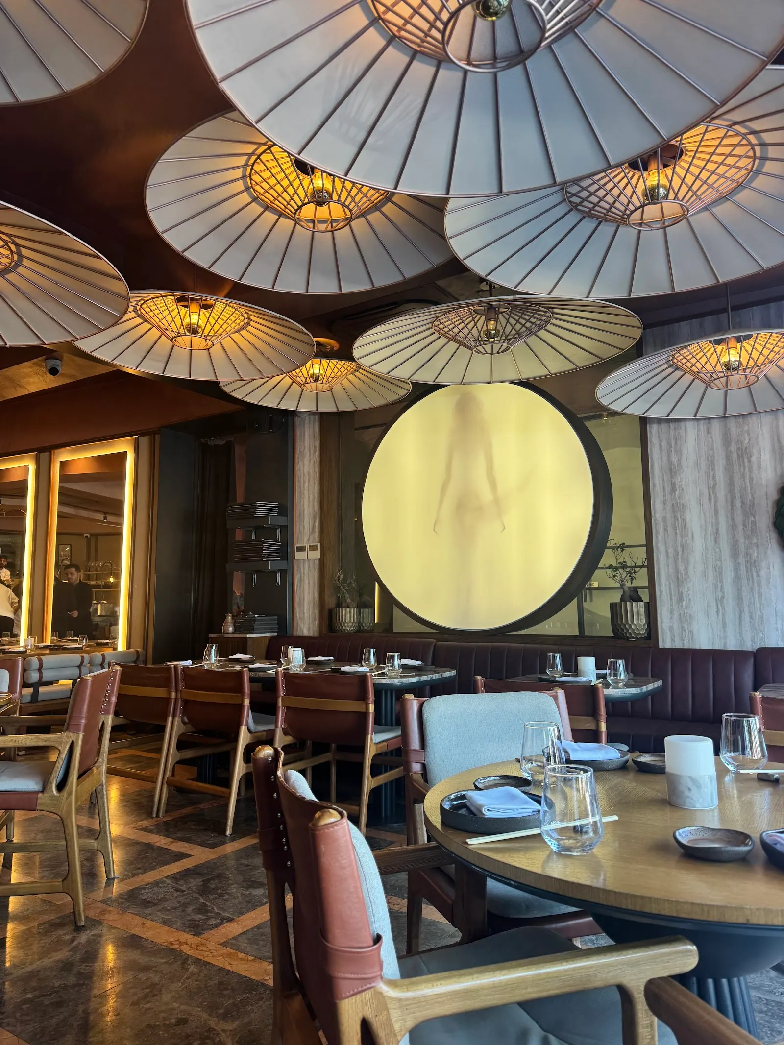 Restaurant interior with patterned ceiling lights, dark wood bar, tables set with place settings, and a large illuminated circular panel on 