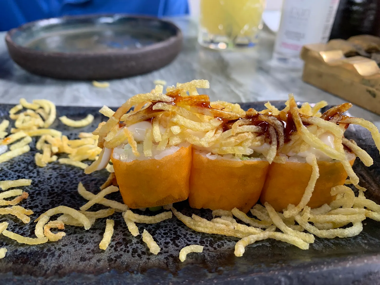 Nigiri sushi topped with crispy element on a decorative black plate with garnish and a small glass beside it.