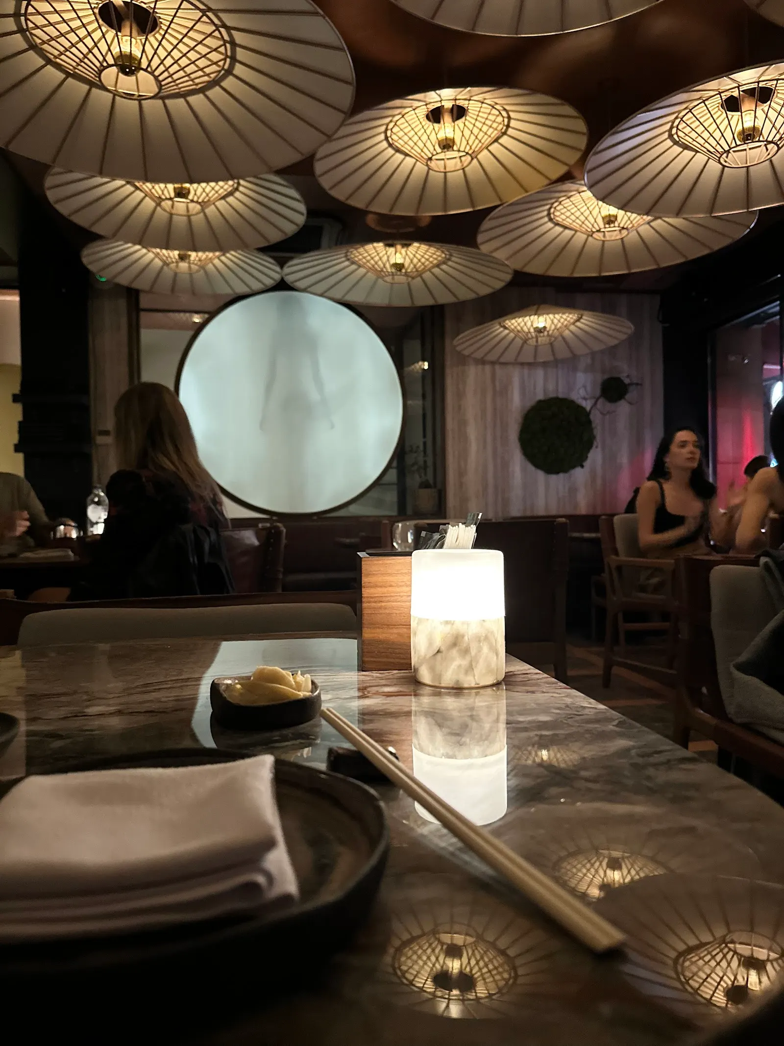 Dining table with place setting and illuminated ceiling feature lights above, a large circular light panel in background, and diners visible