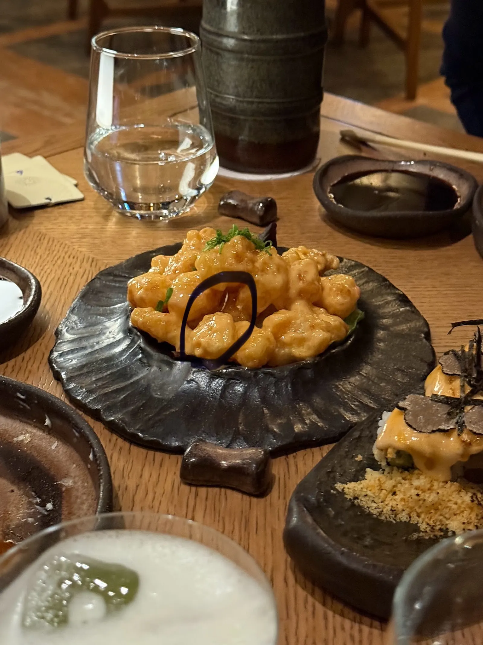 Sushi and fried sushi pieces on dark ceramic plates, glass of water, and small bowls of sauce on a wooden table.
