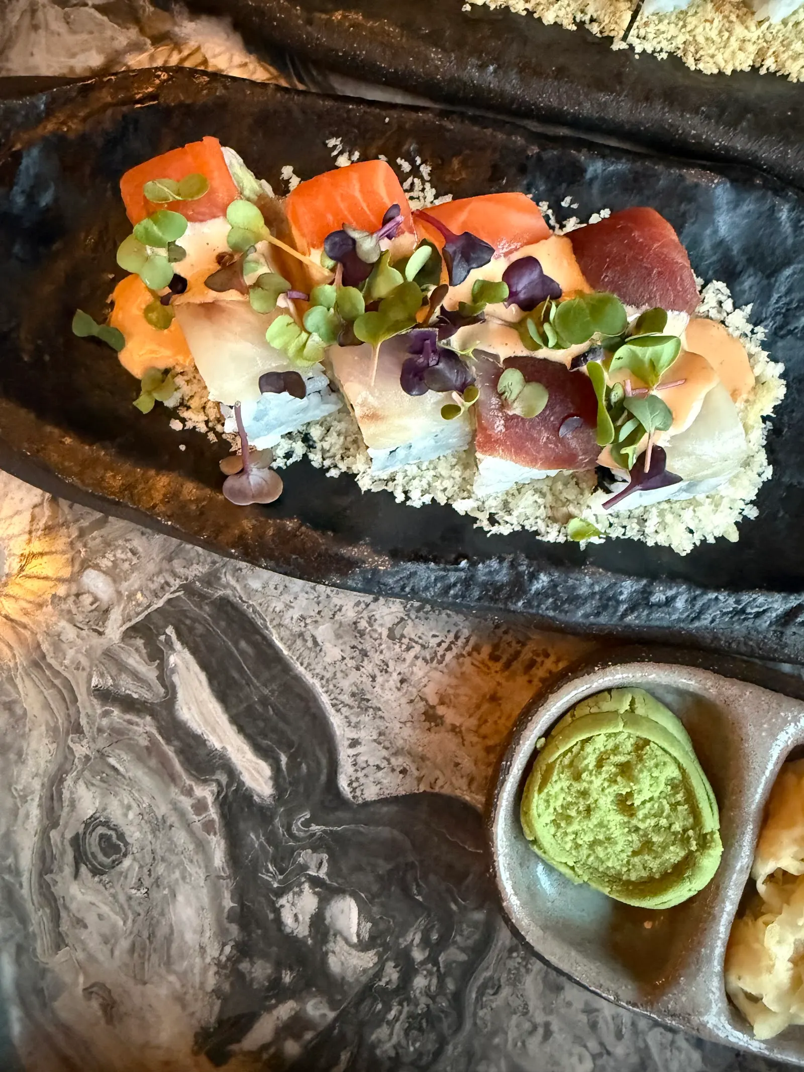 Sashimi slices topped with microgreens and edible flowers on a black stone plate with a small bowl of green sauce.