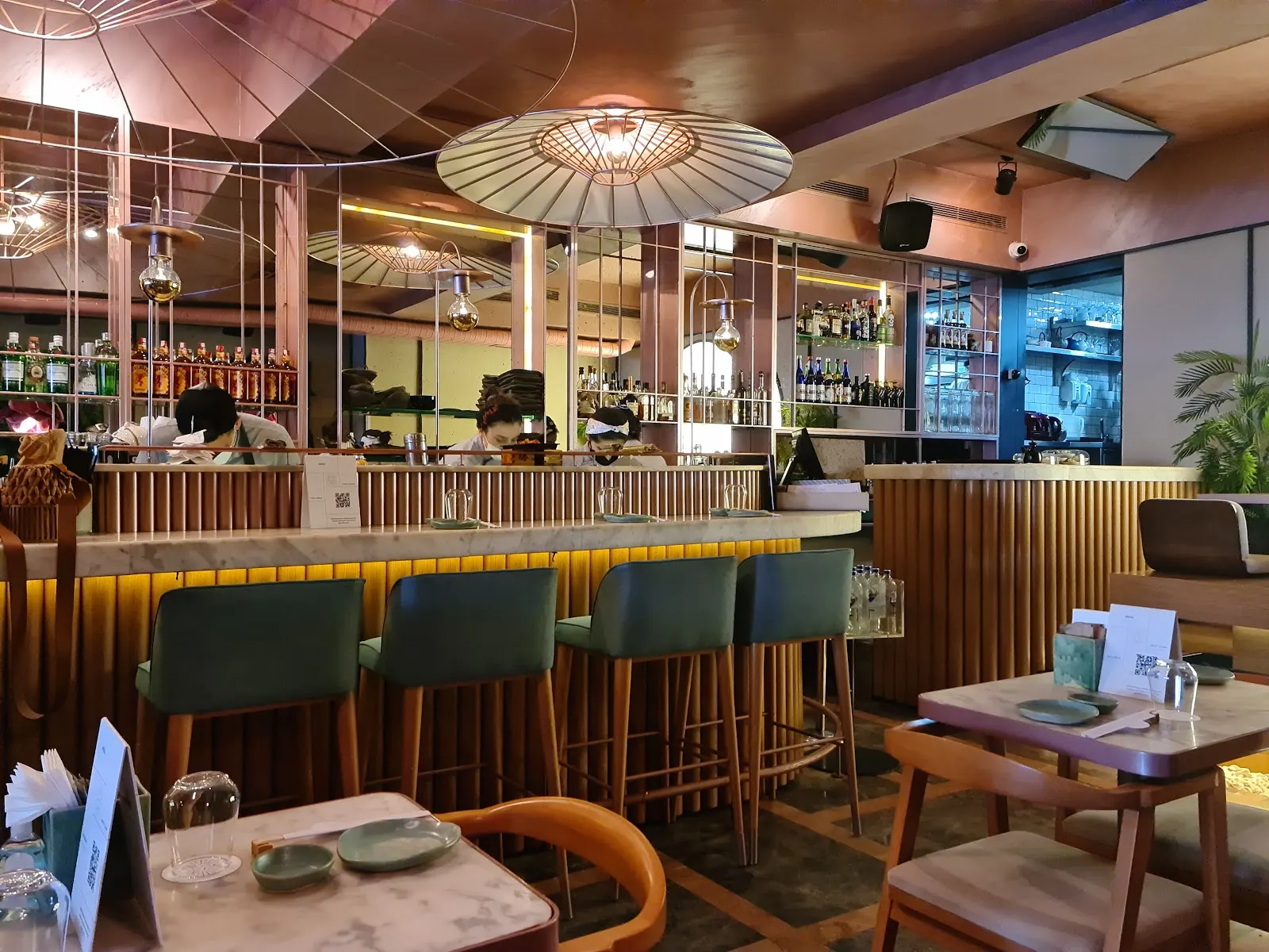 Restaurant dining area with bar seating, green upholstered stools, ceiling feature lights, mirrors, and a table set in the foreground.