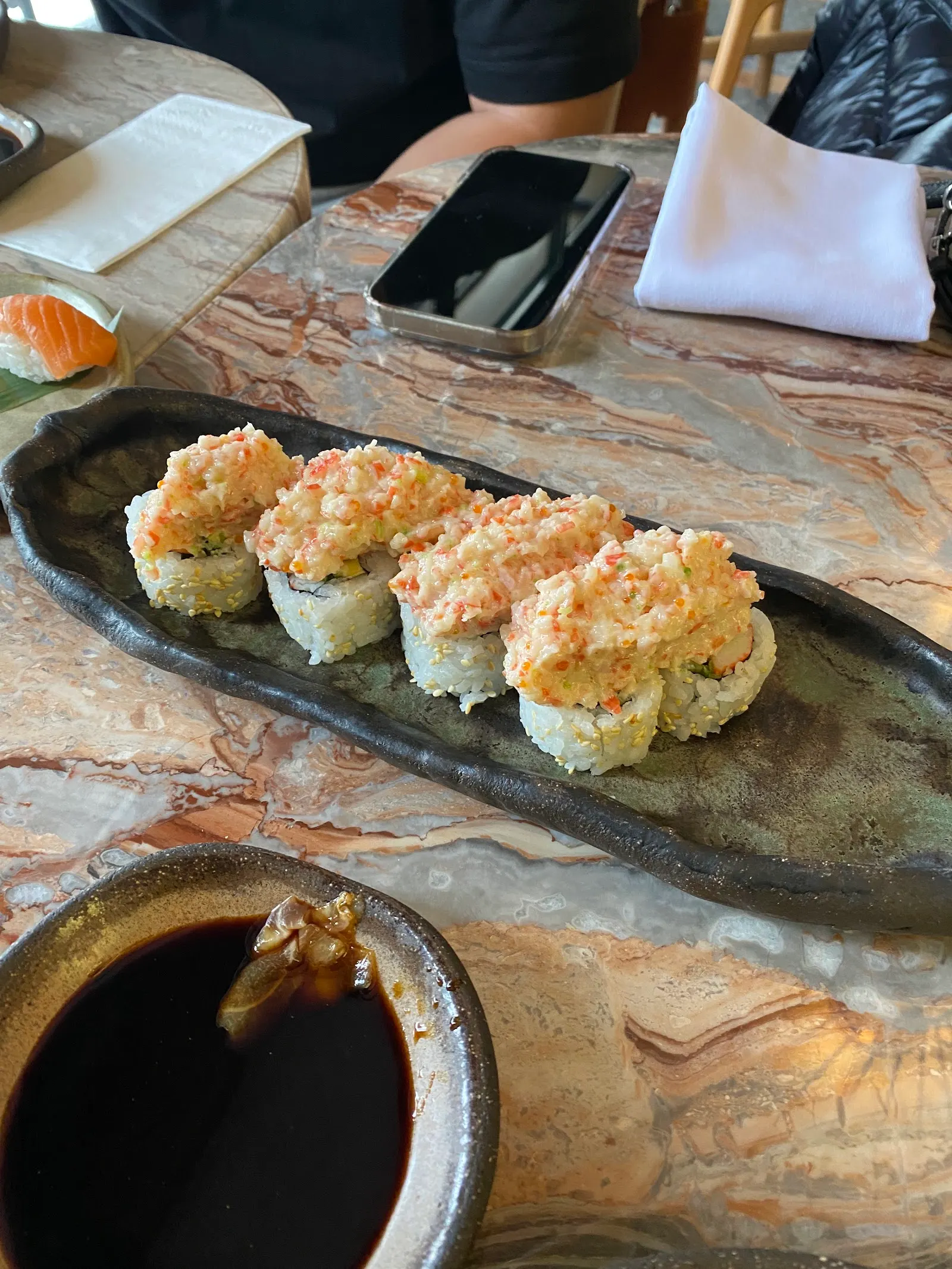 Four pieces of sushi on a dark slate plate with dipping sauce in a small bowl, on a wooden table with diners' hands visible above.